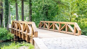 A wooden bridge with a stone pathway leading to it, surrounded by lush greenery and trees.
