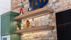 A kitchen with green cabinets, a brick wall, a mounted TV, and a shelf with bottles and bowls.