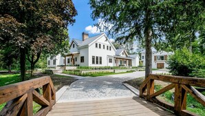 A wooden bridge leading to a white house with a chimney, surrounded by trees and a paved path.