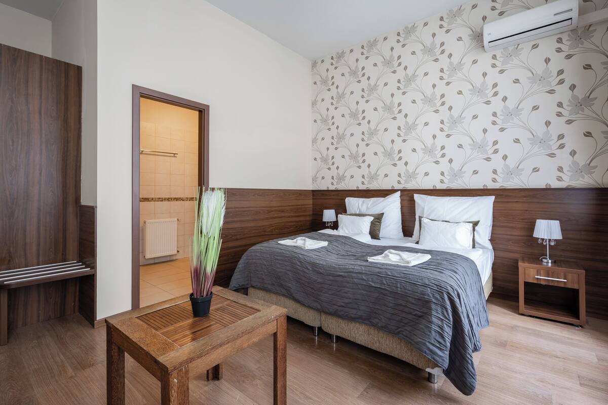 Une chambre d’hôtel avec une tête de lit en bois, un lit avec une literie blanche et grise, une petite table de chevet avec une lampe et une plante en pot.
