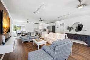 Ein modernes Wohnzimmer mit einer Couch, Sesseln und einem Couchtisch. Es gibt einen Essbereich mit Tisch und Stühlen. Im Hintergrund ist eine Küche mit weißen Schränken und Geräten zu sehen.