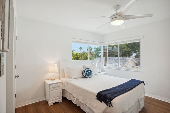 A bedroom with a bed, nightstands, a ceiling fan, and a window with a view of greenery. - Beach Livin 5BR Walk to Beach ~ Rooftop Deck ~3 Kitchens (Treasure Island)