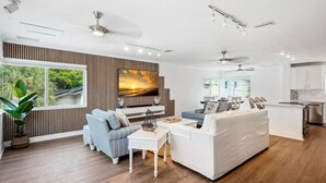 A modern living room with a large TV, a sofa, a coffee table, and a dining area with chairs. - Beach Livin 5BR  Walk to Beach ~ Rooftop Deck ~3 Kitchens (Treasure Island)