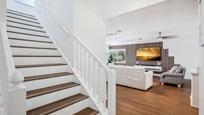 A staircase with wooden steps and white railings leading to a living area with a large TV, a sofa, and a patterned wall. - Beach Livin 5BR  Walk to Beach ~ Rooftop Deck ~3 Kitchens (Treasure Island)