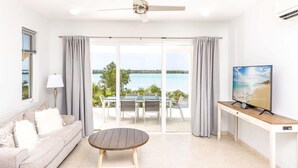 Ein modernes Wohnzimmer mit einer Couch, einem runden Holztisch, einem Deckenventilator, einem Fernseher auf einem Holzschrank und einer großen Schiebeglastür, die zu einem Außenbereich mit Meerblick führt.