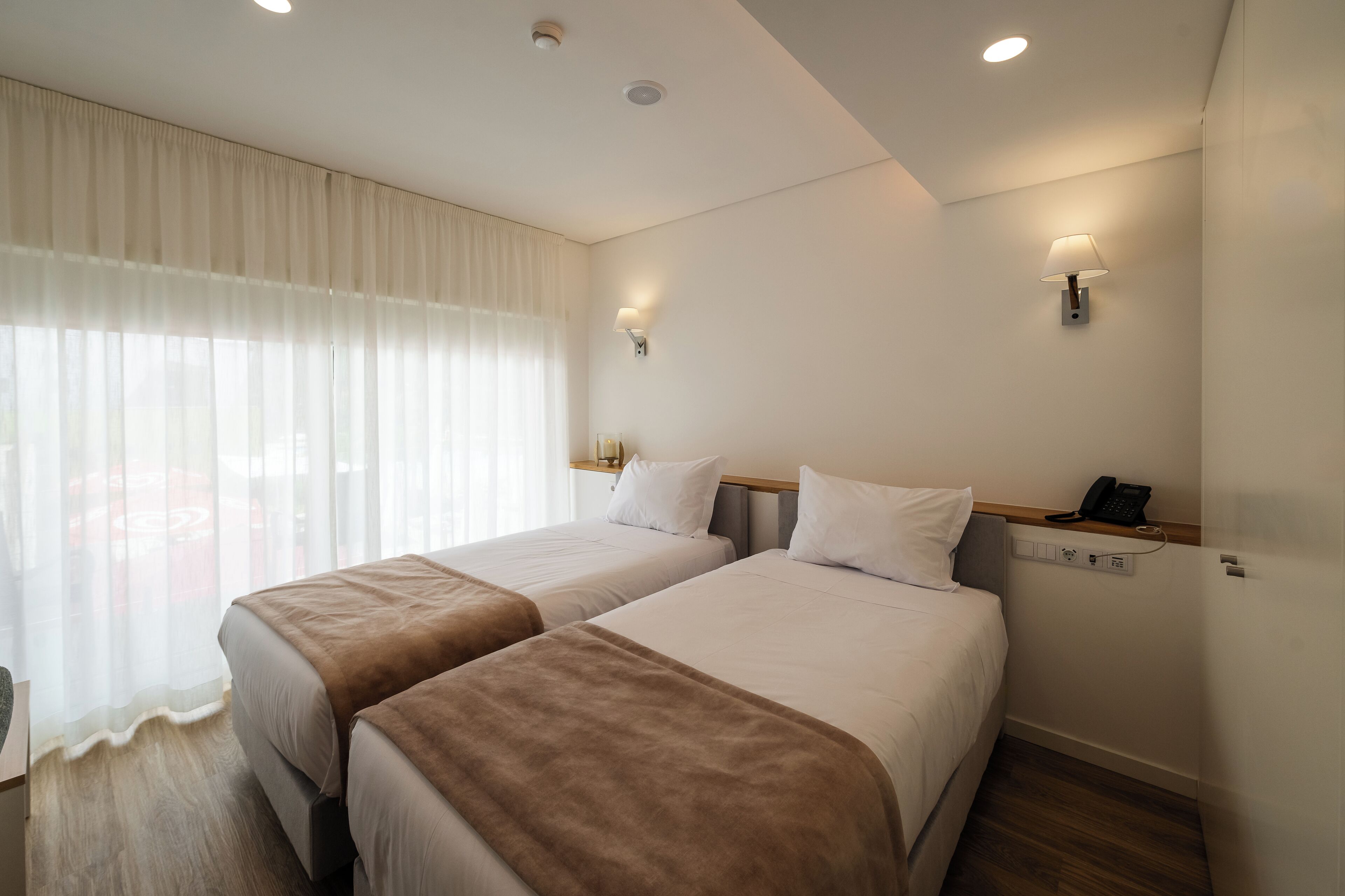Quarto de hotel com duas camas, uma mesa de cabeceira com abajur, um telefone e uma janela com cortinas translúcidas.