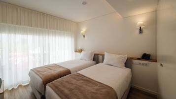 Quarto de hotel com duas camas, uma mesa de cabeceira com abajur, um telefone e uma janela com cortinas translúcidas.