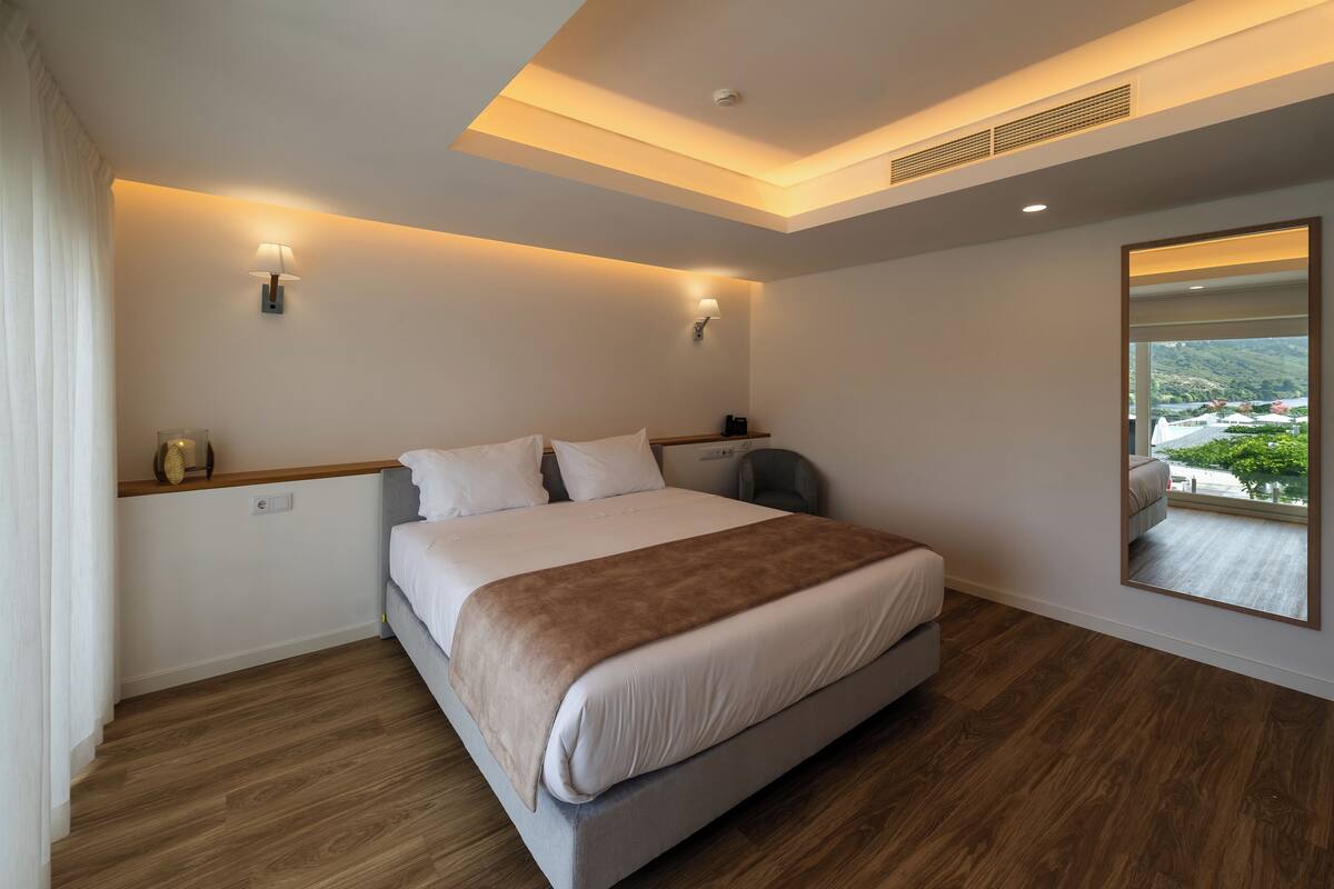 Ein modernes Hotelzimmer mit einem großen Bett, einem Nachttisch, einem Sessel und einem großen Spiegel, der den Raum reflektiert.