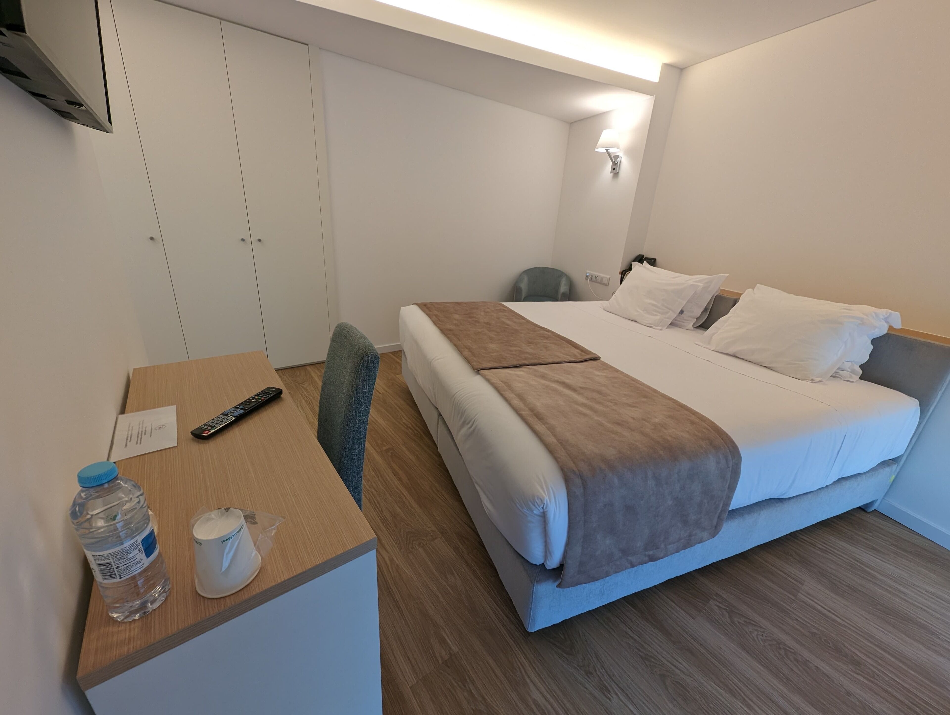A hotel room with a bed, a desk, a chair, a water bottle, a cup, and a remote control.