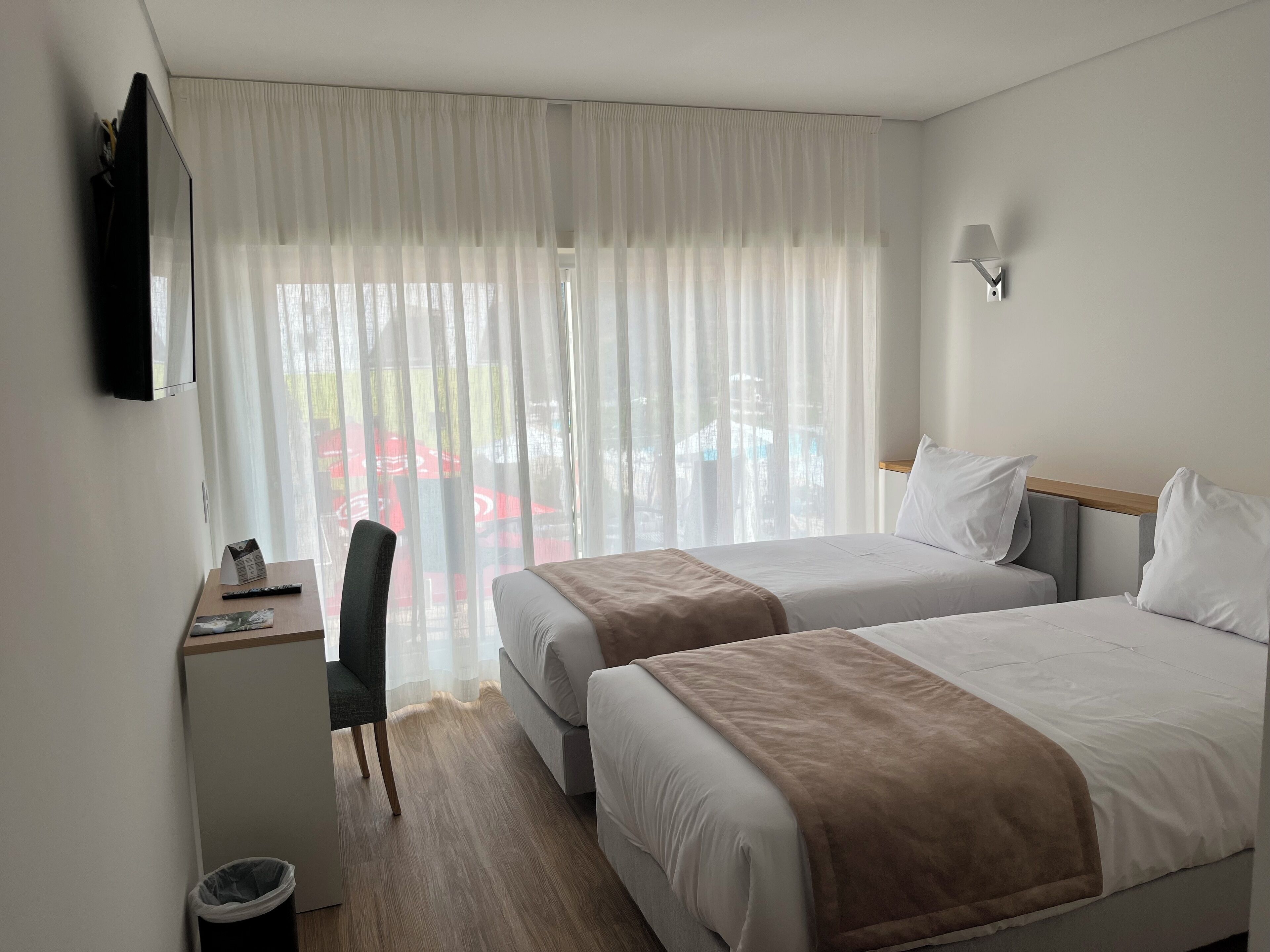 A hotel room with two beds, a desk, a chair, a TV, and a window with sheer curtains.