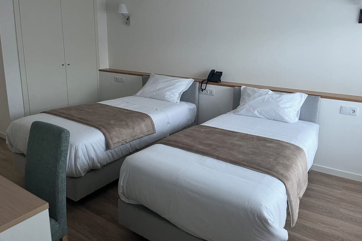 Ein Hotelzimmer mit zwei Einzelbetten, einem Schreibtisch, einem Stuhl, einem Kleiderschrank und einem Telefon an der Wand.