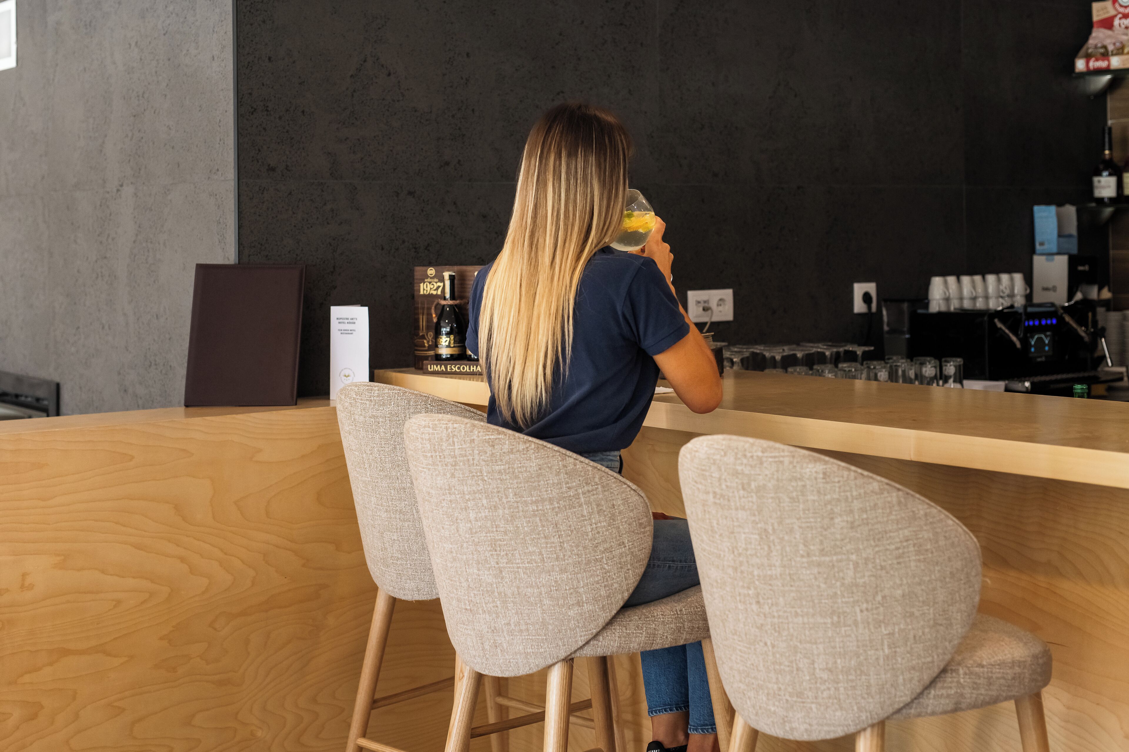 Una persona sentada en un bar, con una bebida en la mano, una máquina de café y un menú al fondo.