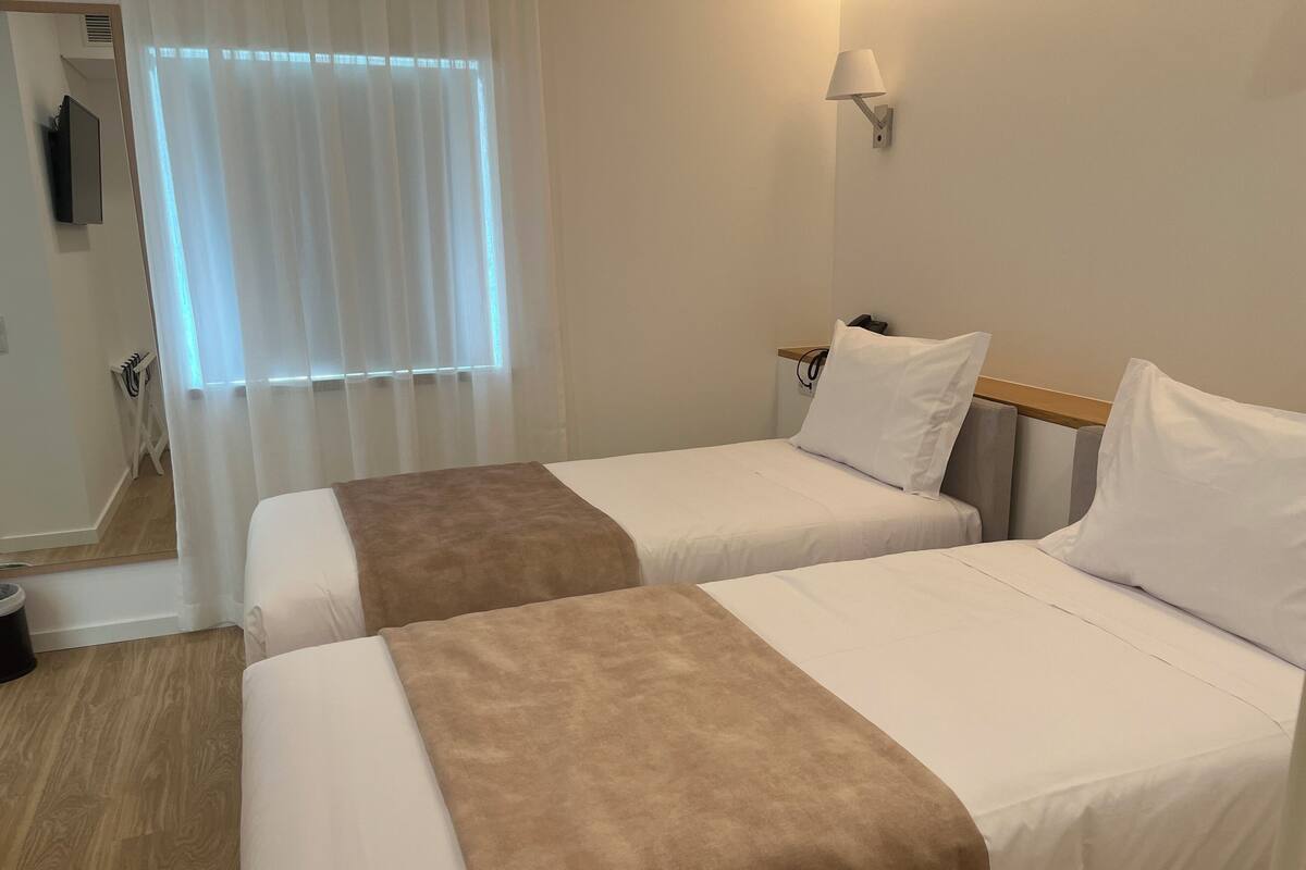 Ein Hotelzimmer mit zwei Betten, einem Fernseher, einer Lampe und einem Fenster mit Vorhängen.