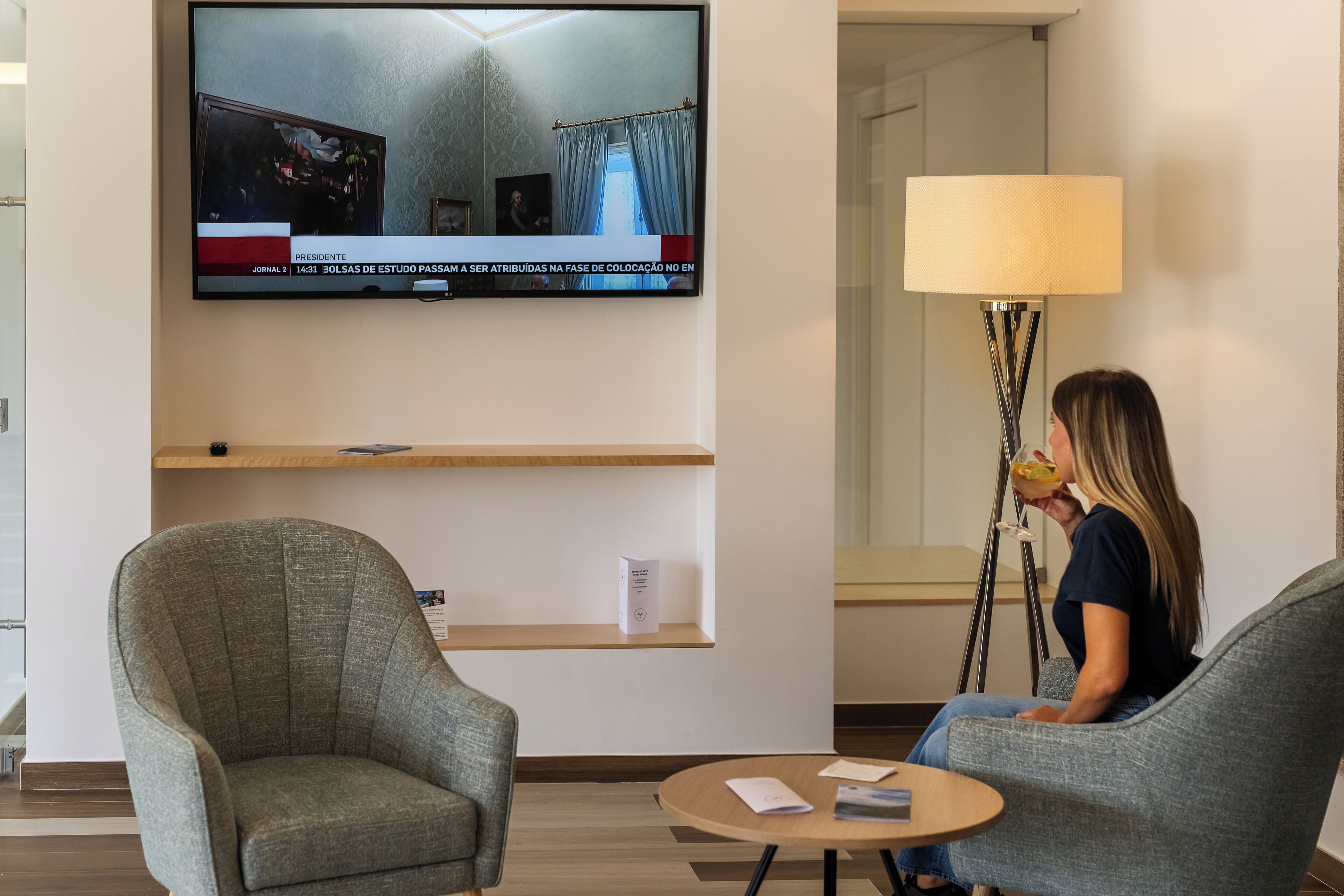 A modern hotel room with a television mounted on the wall displaying a news broadcast, a standing lamp, two upholstered armchairs, and a small round table with a magazine.
