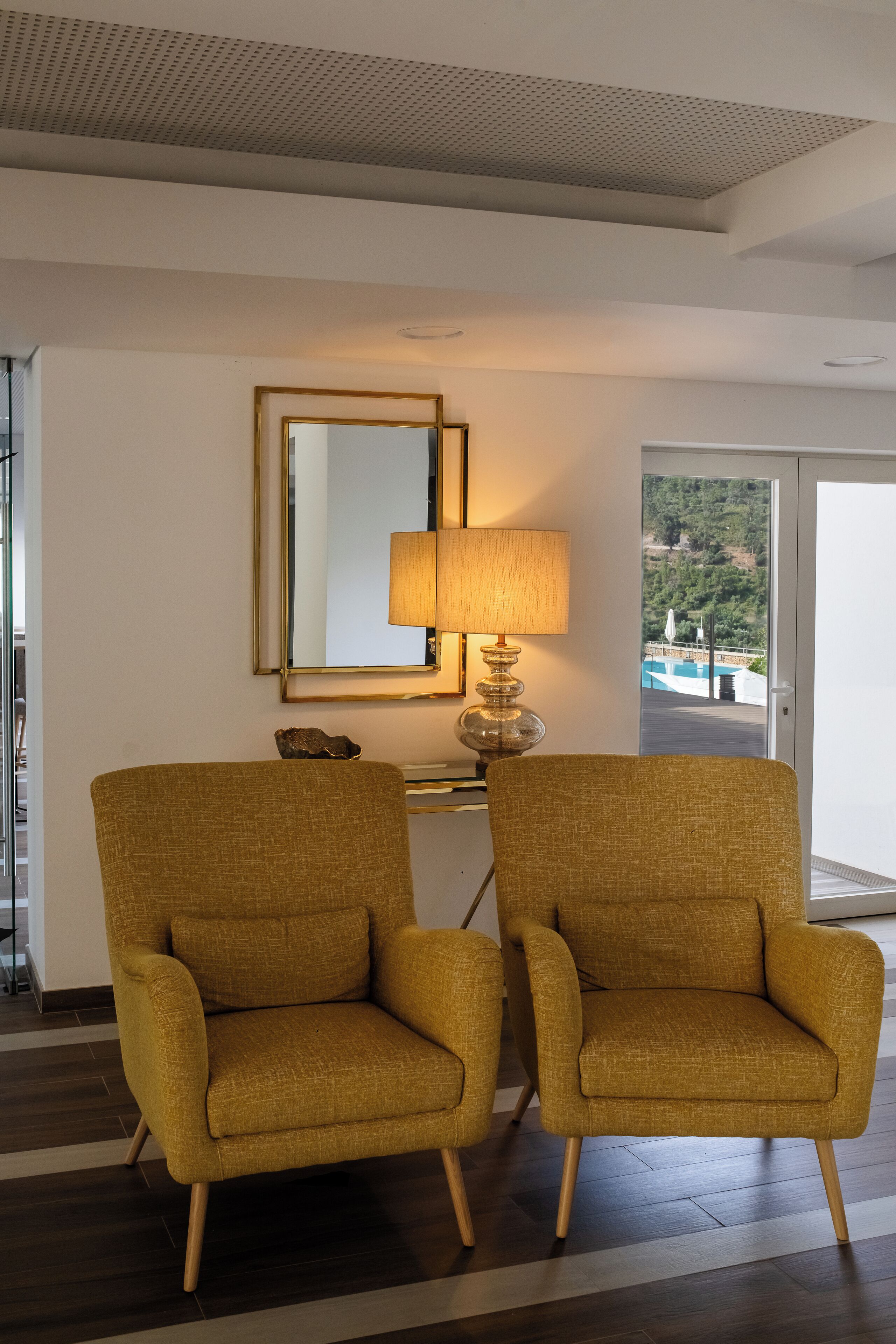 Dos sillones color mostaza con patas de madera, un espejo, una lámpara y vistas a una piscina y follaje a través de una puerta de vidrio.