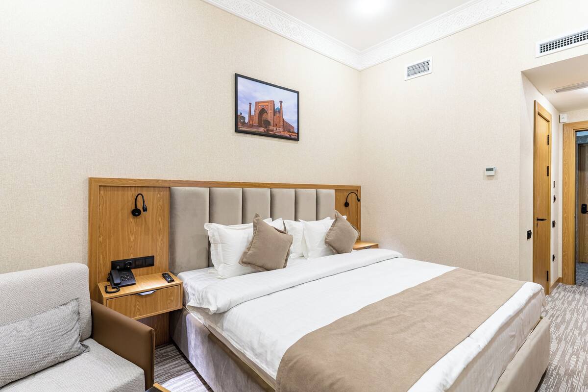 A hotel room with a large bed, a bedside table with a phone and a remote control, a chair, and a picture of a building on the wall.