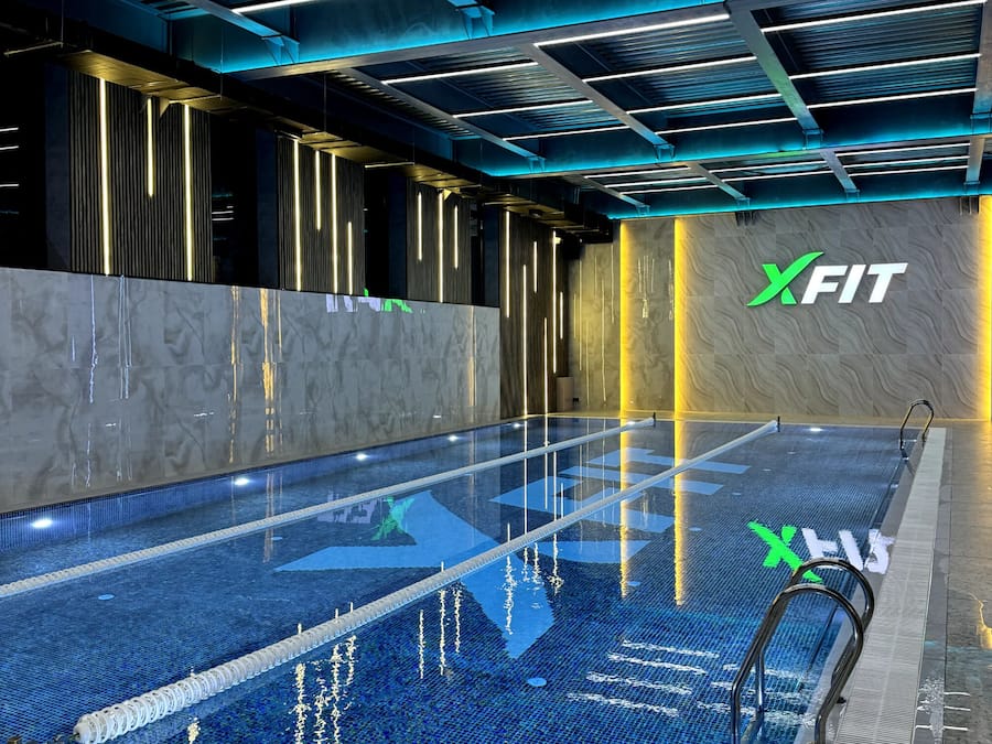 A modern indoor swimming pool with a clear water surface, a tiled floor, and a ceiling with linear lighting fixtures.