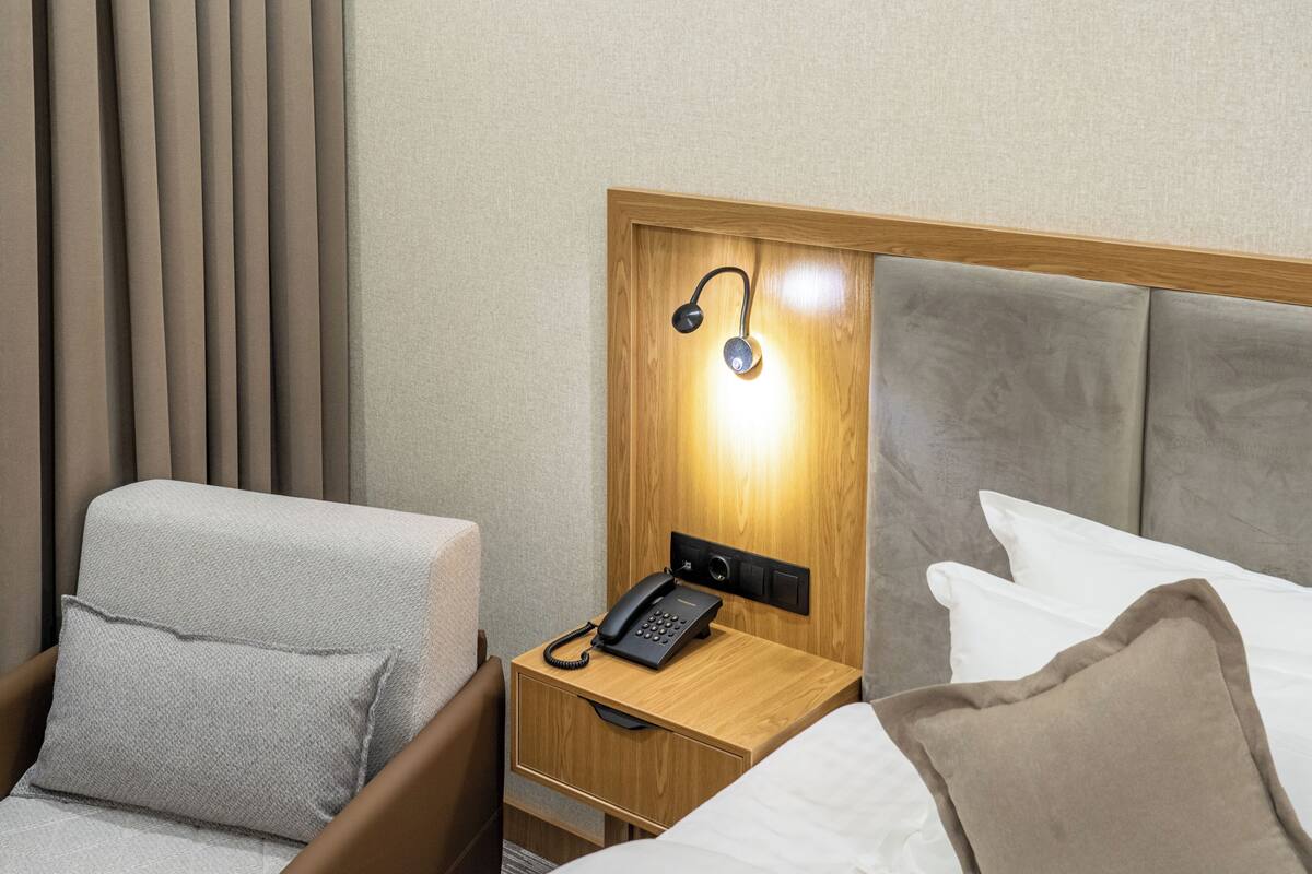 A hotel room with a wooden headboard, a bedside table with a lamp, a telephone, and a bed with pillows.