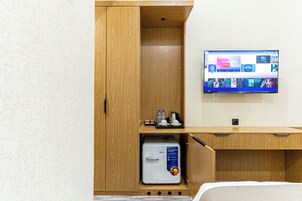A hotel room with a wooden wardrobe, a small refrigerator, a TV mounted on the wall, and a desk with a tray of items.