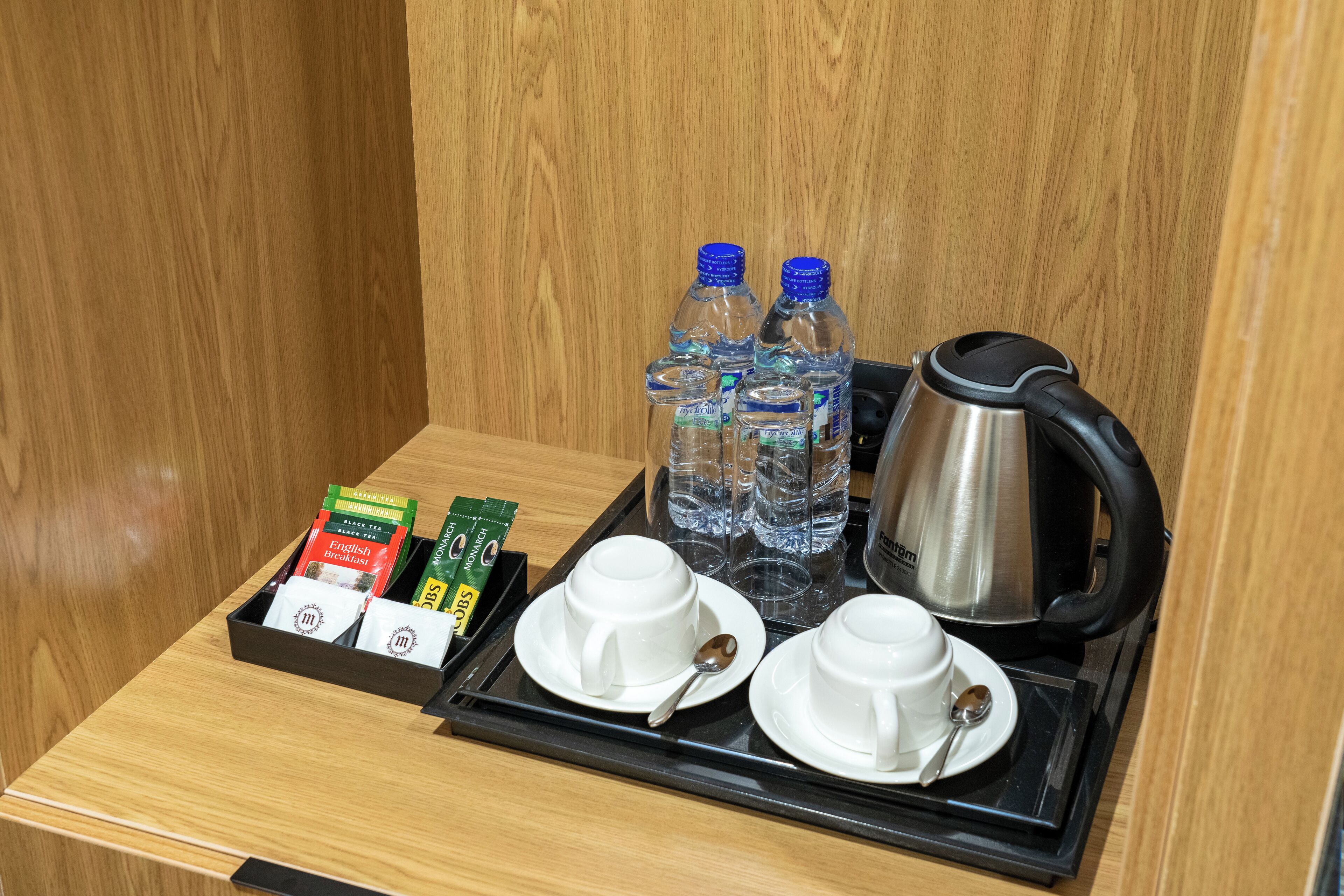 A tray with two white cups, two spoons, two bottles of water, a kettle, and two packets of tea in a wooden room.