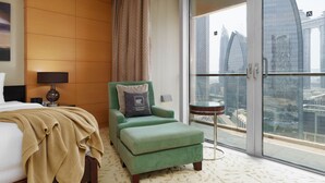 Une chambre d’hôtel avec un lit, un fauteuil vert, un petit pouf, une lampe et une vue sur le paysage urbain, visible à travers une grande fenêtre.