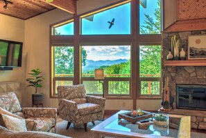 Una sala de estar con ventanas amplias, chimenea de piedra y vistas a montañas.