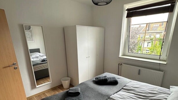 Ett sovrum med en säng, en garderob, en spegel, ett fönster och en hängande lampa.