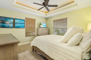Un dormitorio con cama, ventilador de techo, televisor y dos ventanas con persianas.