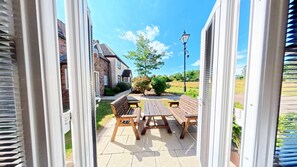 An outdoor patio with wooden benches and tables, a brick house in the background, and a clear blue sky. - Stunning Coastal Cottage - Sleeping 4 in 2 Bedrooms - Pool and Beach Access Included. (Green Close - Near the Front Field)