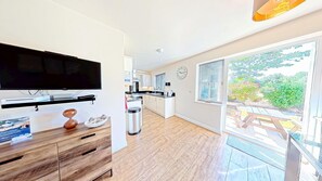 A modern kitchen with a flat-screen TV, wooden cabinetry, and a dining area with a view of greenery. - Stunning Coastal Cottage - Sleeping 4 in 2 Bedrooms - Pool and Beach Access Included. (Green Close - Near the Front Field)