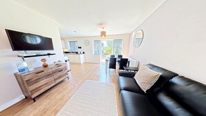 A modern living room with a flat-screen TV, wooden furniture, a black leather sofa, and a dining area with a table and chairs. - Stunning Coastal Cottage - Sleeping 4 in 2 Bedrooms - Pool and Beach Access Included. (Green Close - Near the Front Field)