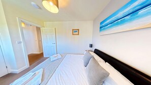 A hotel room with a bed, a painting of a beach, a door, and a light fixture. - Stunning Coastal Cottage - Sleeping 4 in 2 Bedrooms - Pool and Beach Access Included. (Green Close - Near the Front Field)