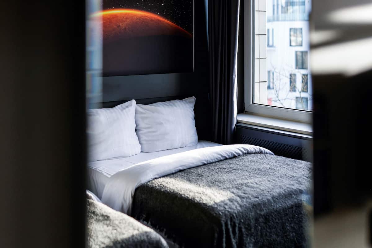 A hotel room with a bed, a window with a view of buildings, and a wall with a space-themed artwork.
