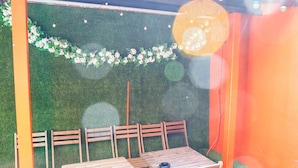 Un espace extérieur aménagé avec des tables et des chaises en bois, un sol recouvert d’un gazon synthétique vert et un mur décoré de fleurs blanches.