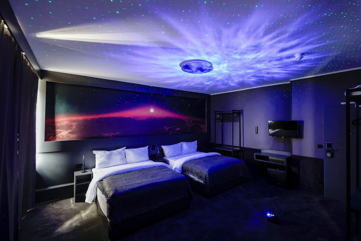A room with two beds, a large wall-mounted TV, and a ceiling with a starry design.