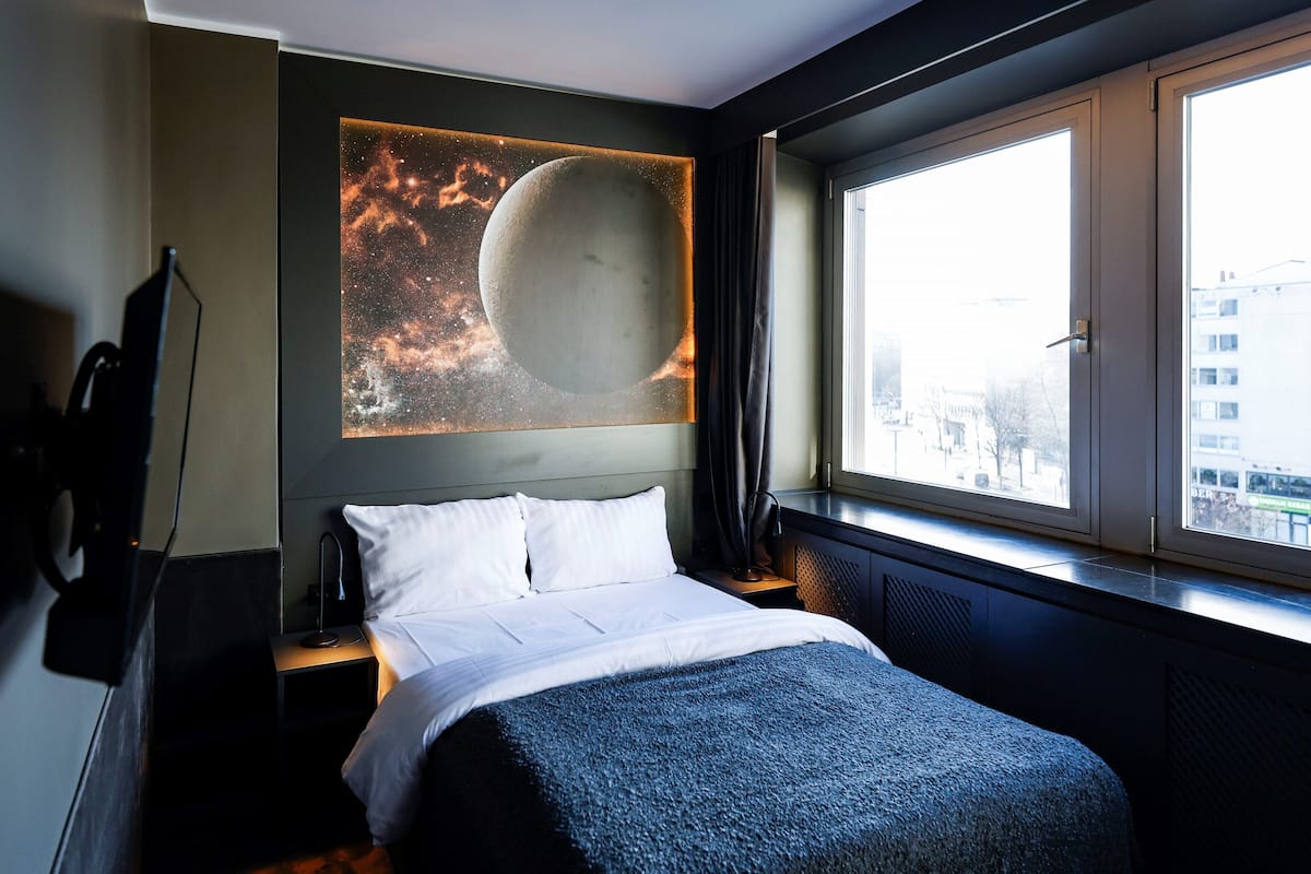A hotel room with a bed, a nightstand, a wall-mounted TV, a window with a view of buildings, and a large artwork of a celestial scene.
