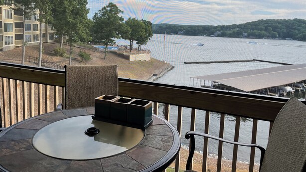 A balcony with a view of a lake, a table with a glass top, and a chair. - Osage Beach Condo getaway! (Osage Beach)