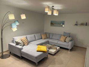 Ein modernes Wohnzimmer mit einem grauen Ecksofa, einem Glastisch und einer Stehlampe mit mehreren Lampenschirmen.