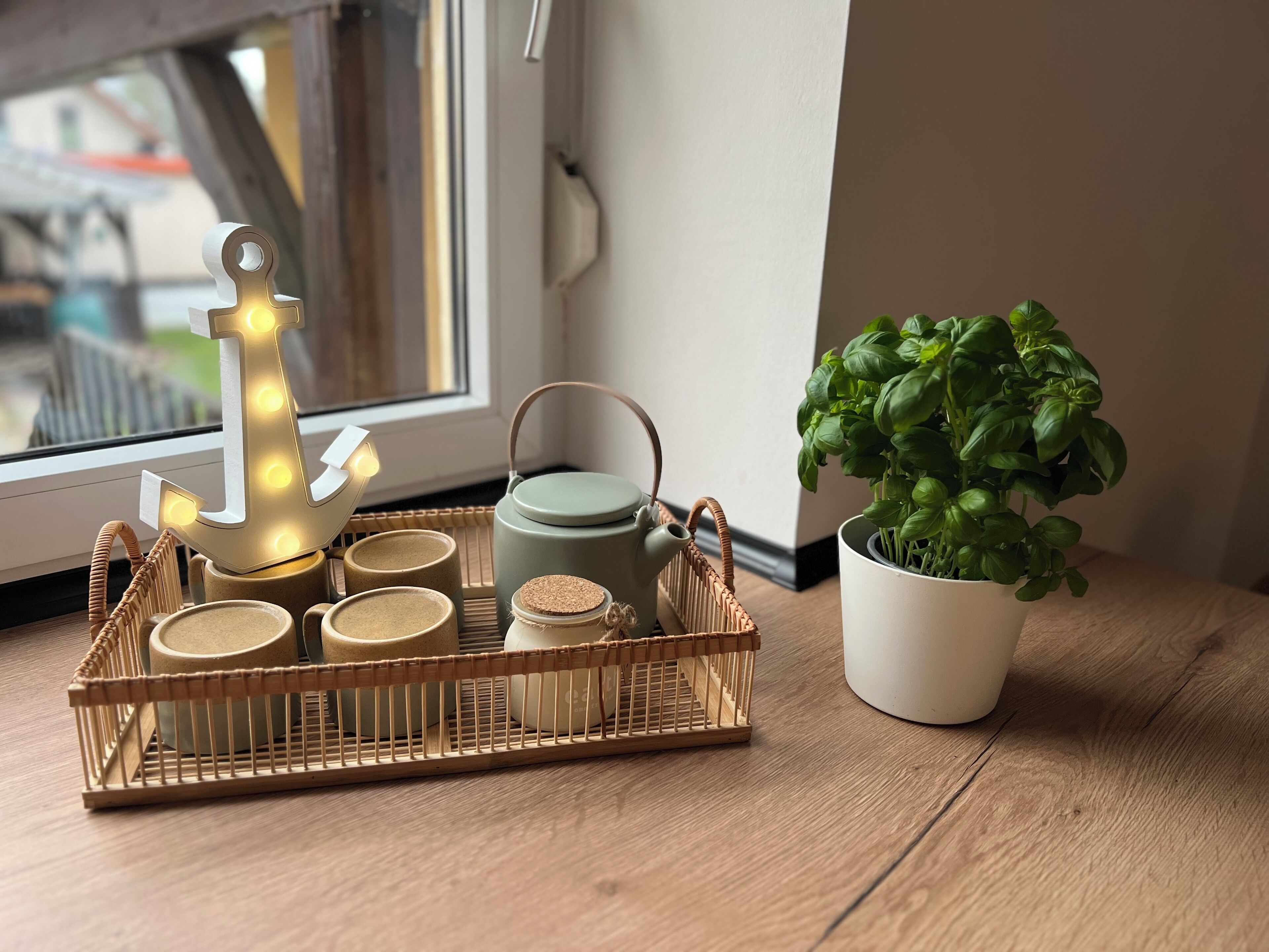 Un plateau avec des tasses à thé, une théière et une plante en pot, posés sur une table en bois près d’une fenêtre.
