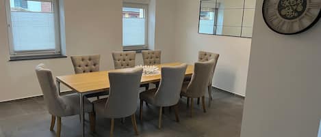 Un comedor con una mesa de madera y seis sillas tapizadas. Encima de la mesa, un candelabro moderno, y a la derecha, un reloj de pared grande.