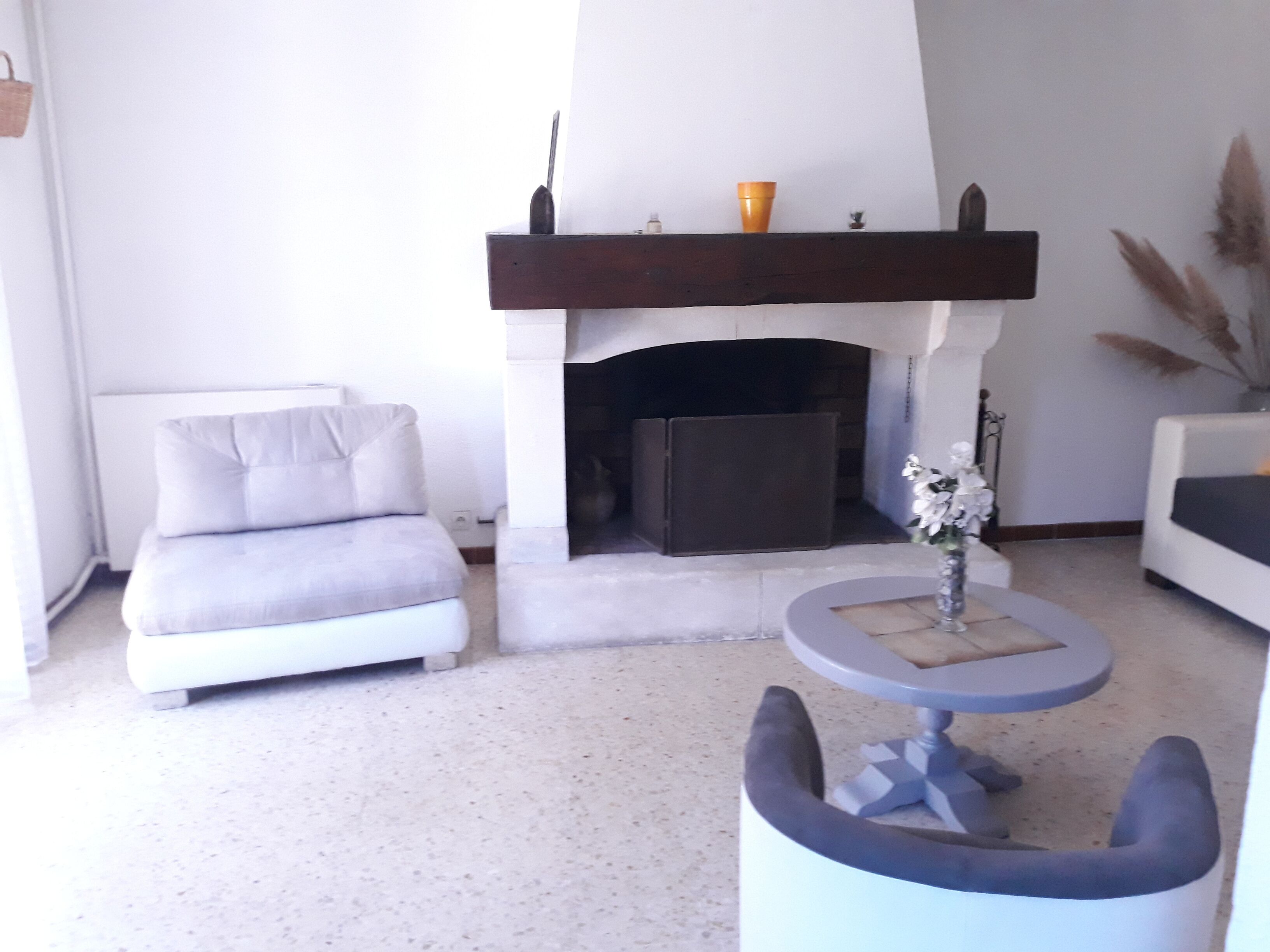 Una sala de estar con chimenea, un sofá gris, una mesa redonda con un florero y un sillón blanco.