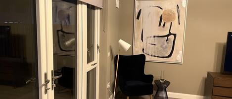 Une pièce dotée d’une porte-fenêtre coulissante, d’une chaise noire, d’une petite table ronde avec une lampe et d’un grand tableau abstrait accroché au mur.
