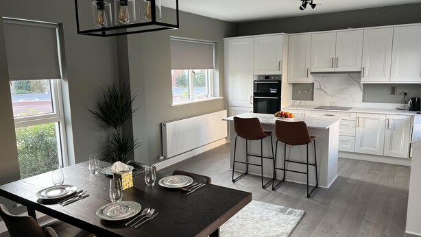 A modern kitchen with white cabinets, a central island with bar stools, and a dining area with a dark wooden table set for four. - Brand New Luxurious and Modern Family House (Lucan)