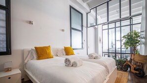 A modern hotel room with a large bed, two pillows, a bedside table, a lamp, and a view of the city through large windows.