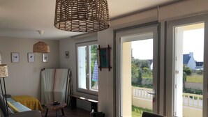 A cozy living room with a sofa, a small table, and a large window with a view of greenery and a white building. - Maison Refaite et Lumineuse, vue Mer, à 5 Minutes de la Plage (Plogoff)
