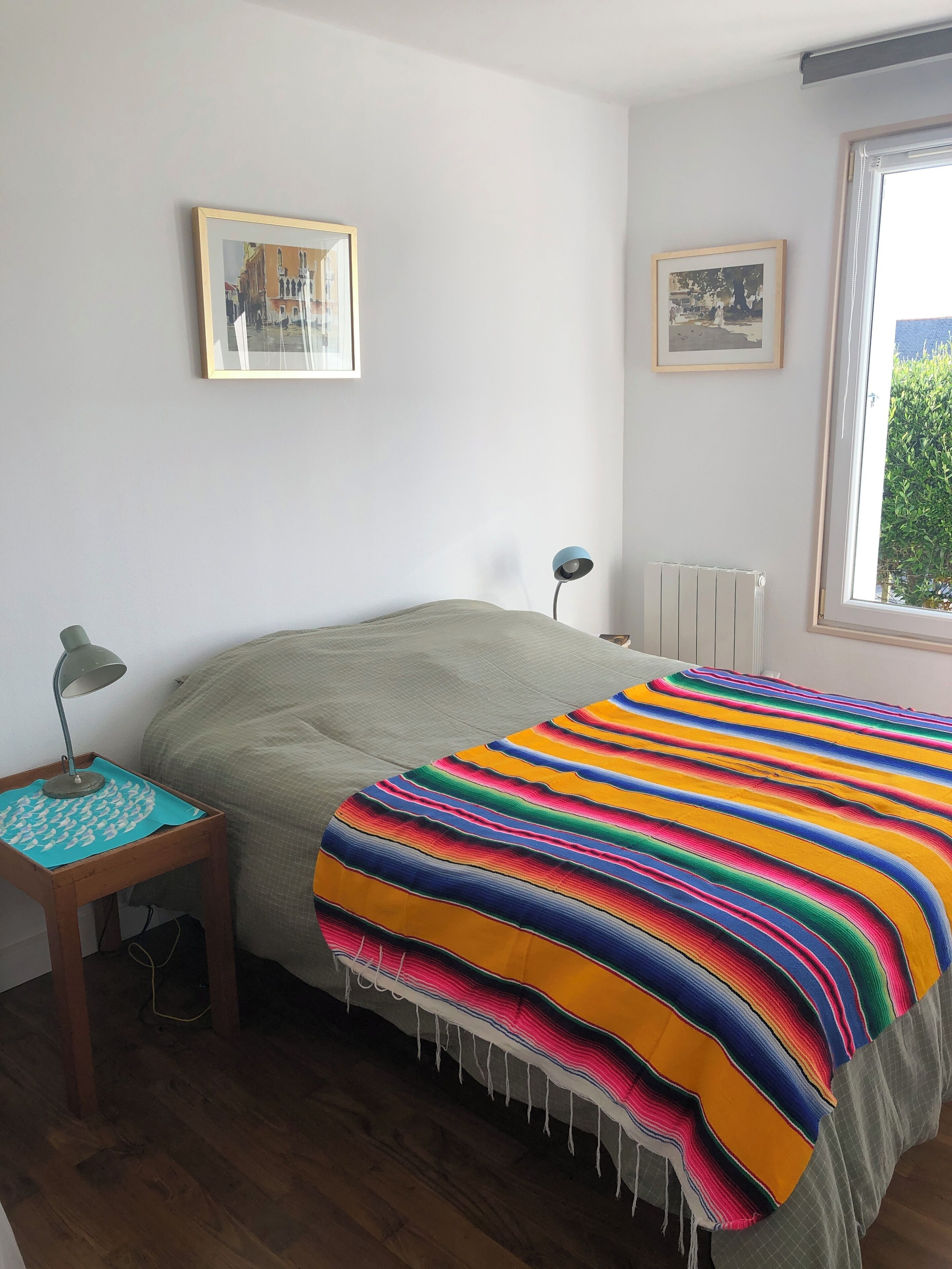 A bedroom with a bed, a nightstand, a lamp, a window, and two framed pictures on the wall.