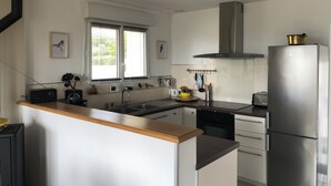 A modern kitchen with white cabinets, a stainless steel refrigerator, a built-in oven, and a window with blinds. - Maison Refaite et Lumineuse, vue Mer, à 5 Minutes de la Plage (Plogoff)