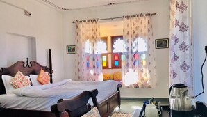 A bedroom with a wooden bed, colorful pillows, a coffee table with a kettle, and curtains with a floral pattern. - Deogarh Hills A Village Experience Property (Deogarh Madaria)