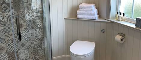 A bathroom with a glass shower enclosure, a toilet, a window with a view, and a shelf with towels and toiletries.