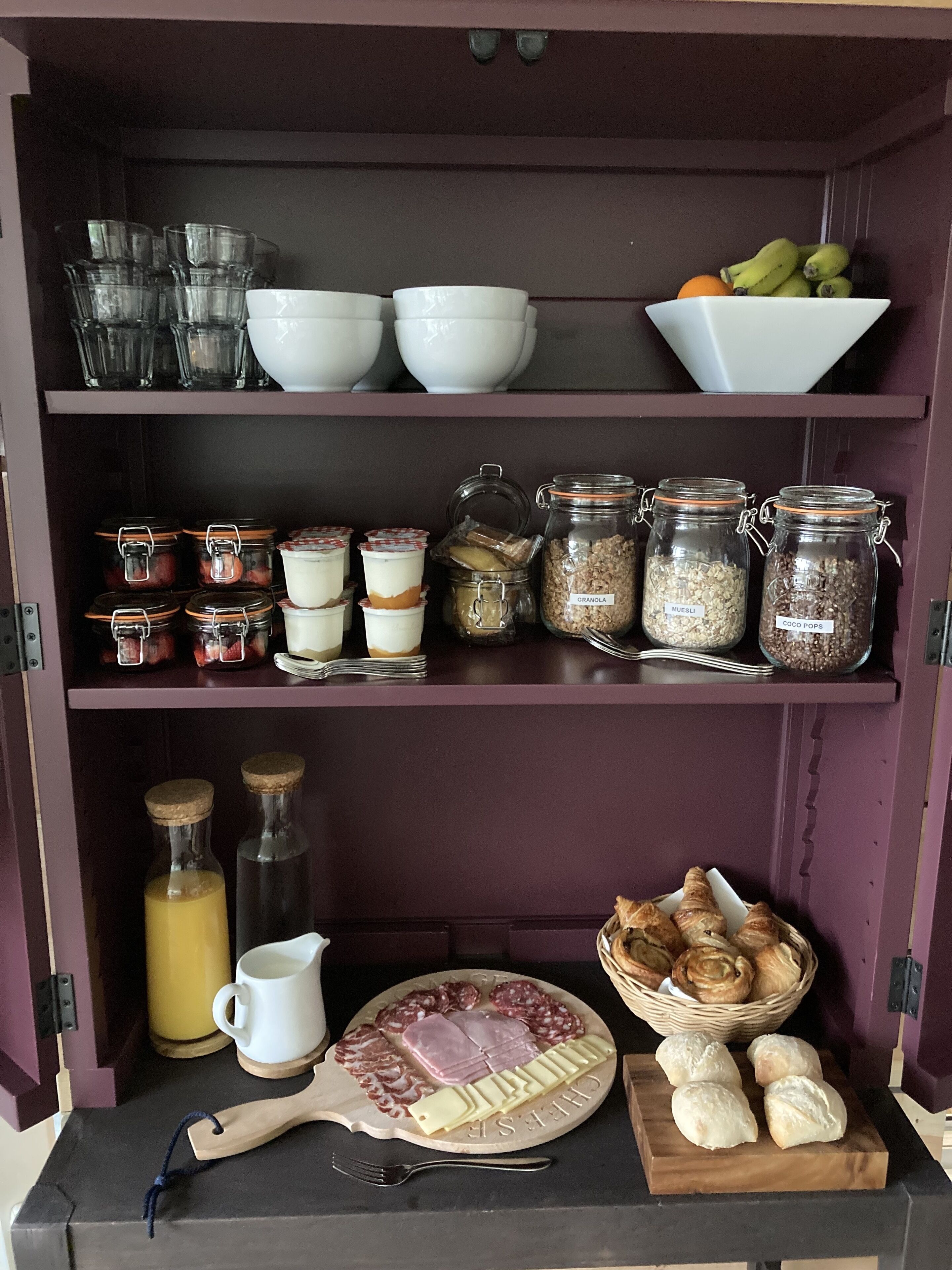 Uma despensa com potes de vidro etiquetados com alimentos, um prato de queijos e frios, e uma tigela de frutas.